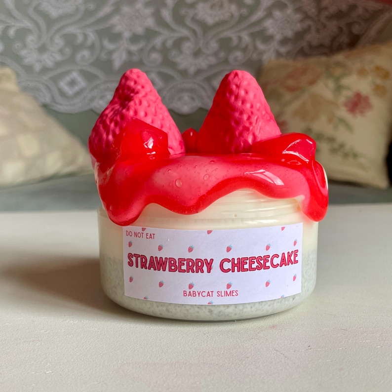 A container labeled "strawberry cheesecake by babycat slimes" is filled with a creme coloured slime with a sand crust, topped with red slime with jelly cubes and strawberry clay pieces.