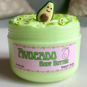 Peut inclure: Un pot de beurre corporel vert clair sur le thème de l'avocat. Il est orné de paillettes en forme d'avocat et d'une tranche d'avocat souriante. L'étiquette rose porte les mots "Avocado Body Butter" et "Do Not Eat".
