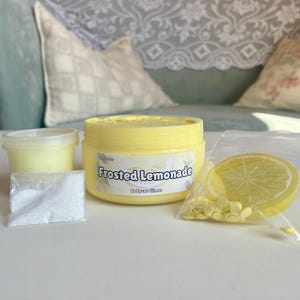 Peut inclure: Un pot de slime jaune avec une &eacute;tiquette qui indique "Frosted Lemonade" et "Babycat Slimes". Le pot est entour&eacute; de poudre blanche, de paillettes jaunes et d'une tranche de citron jaune.