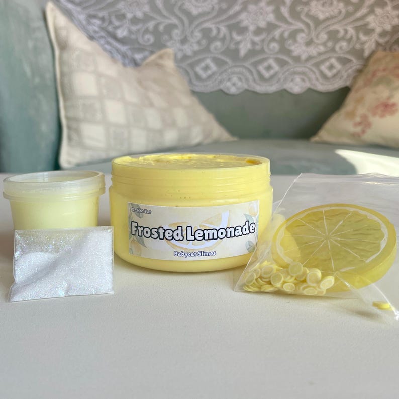 Peut inclure: Un pot de slime jaune avec une &eacute;tiquette qui indique "Frosted Lemonade" et "Babycat Slimes". Le pot est entour&eacute; de poudre blanche, de paillettes jaunes et d'une tranche de citron jaune.