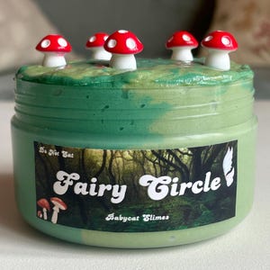 May include: A jar of green slime with five red and white mushroom decorations on top. The jar has a label that reads "Fairy Circle" with a forest scene and the words "Babycat Slimes". The words "Do Not Eat" are also visible.