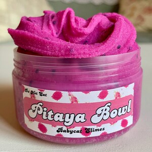 A round transparent container is full of magenta slime with tiny black seeds, resembling a pitaya dragonfruit bowl. on top are beeswax "coconut flakes" and fruit slice charms.