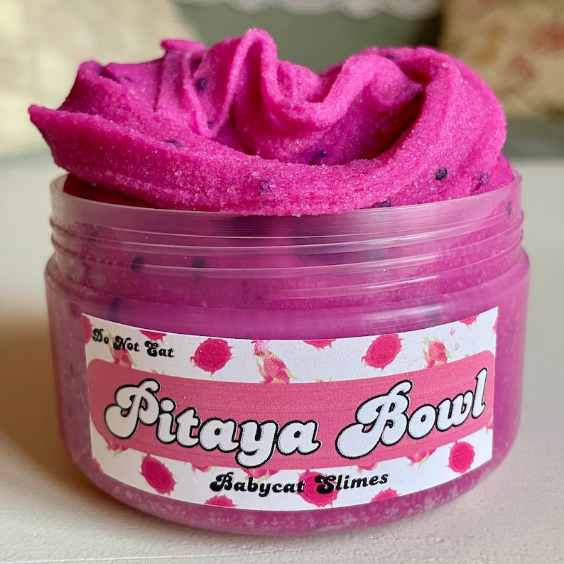 A round transparent container is full of magenta slime with tiny black seeds, resembling a pitaya dragonfruit bowl. on top are beeswax "coconut flakes" and fruit slice charms.