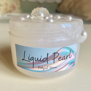 Op de afbeelding: Een kleine, doorzichtige plastic pot met "Liquid Pearl" slijm, met een parelmoerglans. De deksel is versierd met kleine, witte parelachtige kralen. Het etiket bevat de tekst "Liquid Pearl" en "Babycat Slimes" in een cursief lettertype.