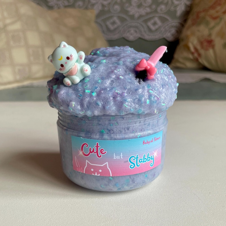 May include: A jar of purple slime with iridescent glitter. A small, light blue cat figurine sits on top. The jar has a label that reads "Cute but Stabby" with a cat graphic. A pink bow and knife are also in the slime.