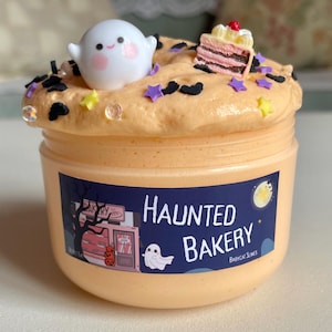 Könnte beinhalten: Ein Glas orangefarbener Schleim mit Halloween-Motiv. Der Schleim ist mit einer weißen Geisterfigur, stern- und fledermausförmigen Streuseln und einem Miniatur-Kuchenstück verziert. Das Glas trägt die Aufschrift "Haunted Bakery".