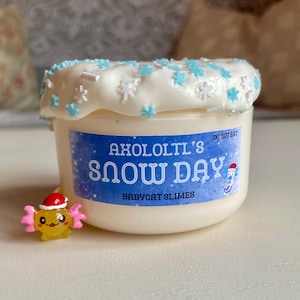 Peut inclure: Un petit pot blanc de slime avec une étiquette bleue et blanche indiquant "Axololt's Snow Day" et "Babycat Slimes". Le slime est garni d'un glaçage blanc et de paillettes flocons de neige bleues et blanches. Une petite figurine d'axolotl jaune avec un bonnet de Père Noël est à côté.