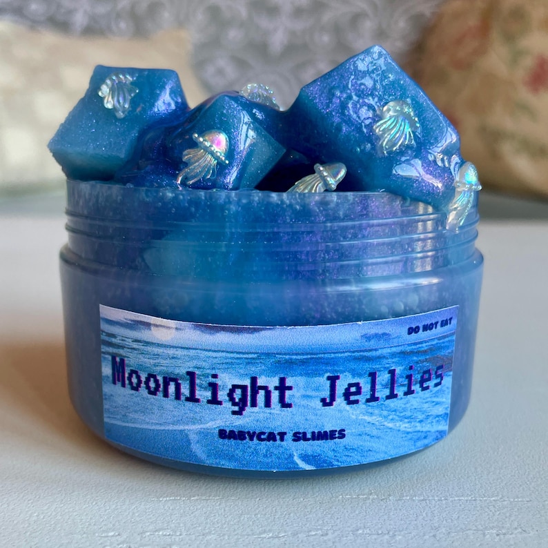 A clear, round plastic container is overflowing with blue sparkly jelly cube slime. The slime is a clear coated slime colorshifting from blue to purple, and has big squishable jelly cubes. It also has tiny opalescent jellyfish charms.