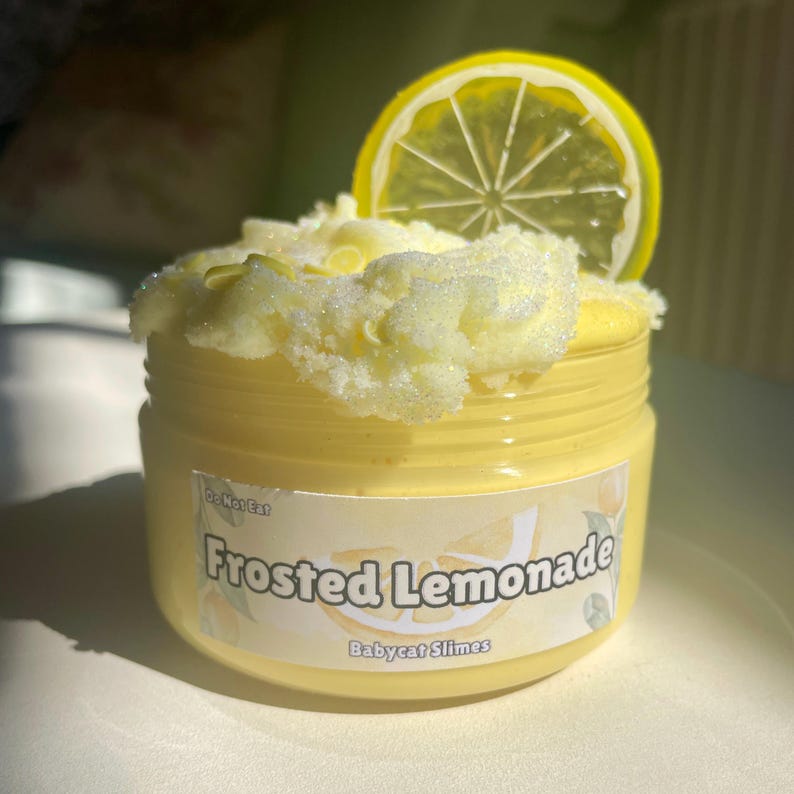 Peut inclure: Une slime jaune dans un pot transparent avec une &eacute;tiquette blanche qui dit "Frosted Lemonade" et "Babycat Slimes". Une tranche de citron jaune est sur le dessus de la slime.