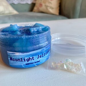 A clear, round plastic container is overflowing with blue sparkly jelly cube slime. The slime is a clear coated slime colorshifting from blue to purple, and has big squishable jelly cubes. It also has tiny opalescent jellyfish charms.