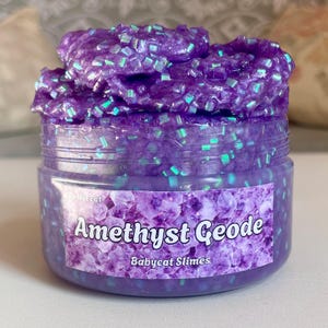 Könnte beinhalten: Lilafarbener Schleim mit schimmerndem Glitzer in einem durchsichtigen Glas mit einem Etikett, auf dem "Amethyst Geode" und "Babycat Slimes" steht. Nicht essen.