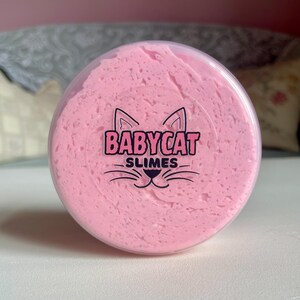 A container labeled "strawberry cheesecake by babycat slimes" is filled with a creme coloured slime with a sand crust, topped with red slime with jelly cubes and strawberry clay pieces.