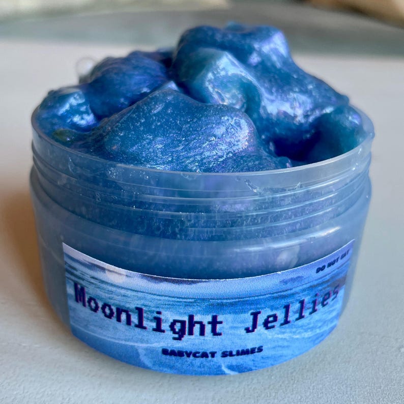 A clear, round plastic container is overflowing with blue sparkly jelly cube slime. The slime is a clear coated slime colorshifting from blue to purple, and has big squishable jelly cubes. It also has tiny opalescent jellyfish charms.