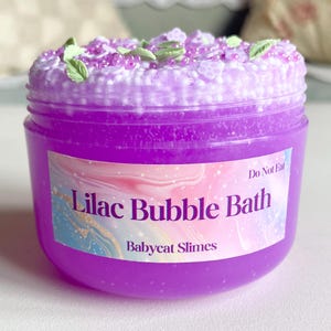May include: A jar of purple Lilac Bubble Bath by Babycat Slimes. The jar has a label with the product name and brand. The top of the bath product is decorated with white and purple beads and green leaf accents. The label also says "Do Not Eat".
