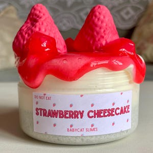 A container labeled "strawberry cheesecake by babycat slimes" is filled with a creme coloured slime with a sand crust, topped with red slime with jelly cubes and strawberry clay pieces.
