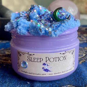 May include: A jar of lavender Sleep Potion with a blue, cloud-like topping and star-shaped glitter. A green crescent moon sits on top. The label reads "Sleep Potion" with the text "Starlight and lavender mist - may slumber find you swift."