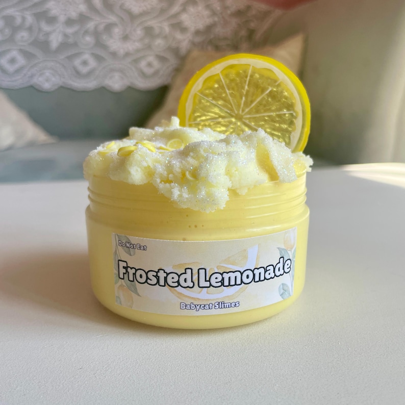 Peut inclure: Une slime jaune dans un pot transparent avec une tranche de citron jaune sur le dessus. Le pot a une &eacute;tiquette blanche qui dit "Frosted Lemonade" et "Babycat Slimes".