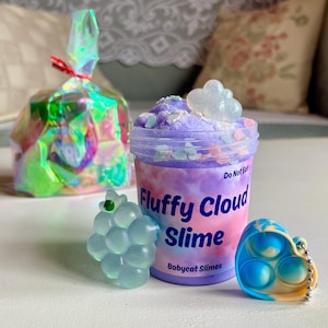 Op de afbeelding: Een verzameling kleurrijk sensorisch speelgoed. Bevat een pot "Fluffy Cloud Slime" met een wolk-vormige bedel, een blauw en wit hartvormig pop-it speelgoed, een doorzichtige druif-vormige bedel en een cellofaan zakje vastgebonden met een rood lint.