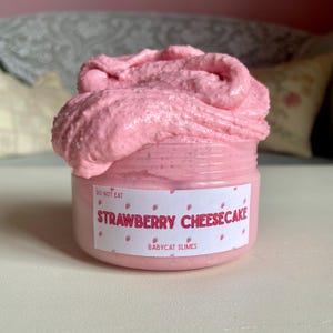 A container labeled "strawberry cheesecake by babycat slimes" is filled with a creme coloured slime with a sand crust, topped with red slime with jelly cubes and strawberry clay pieces.