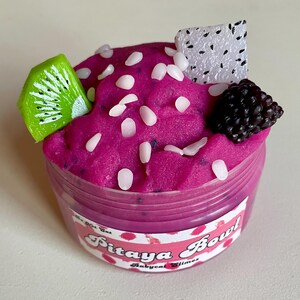 A round transparent container is full of magenta slime with tiny black seeds, resembling a pitaya dragonfruit bowl. on top are beeswax "coconut flakes" and fruit slice charms.