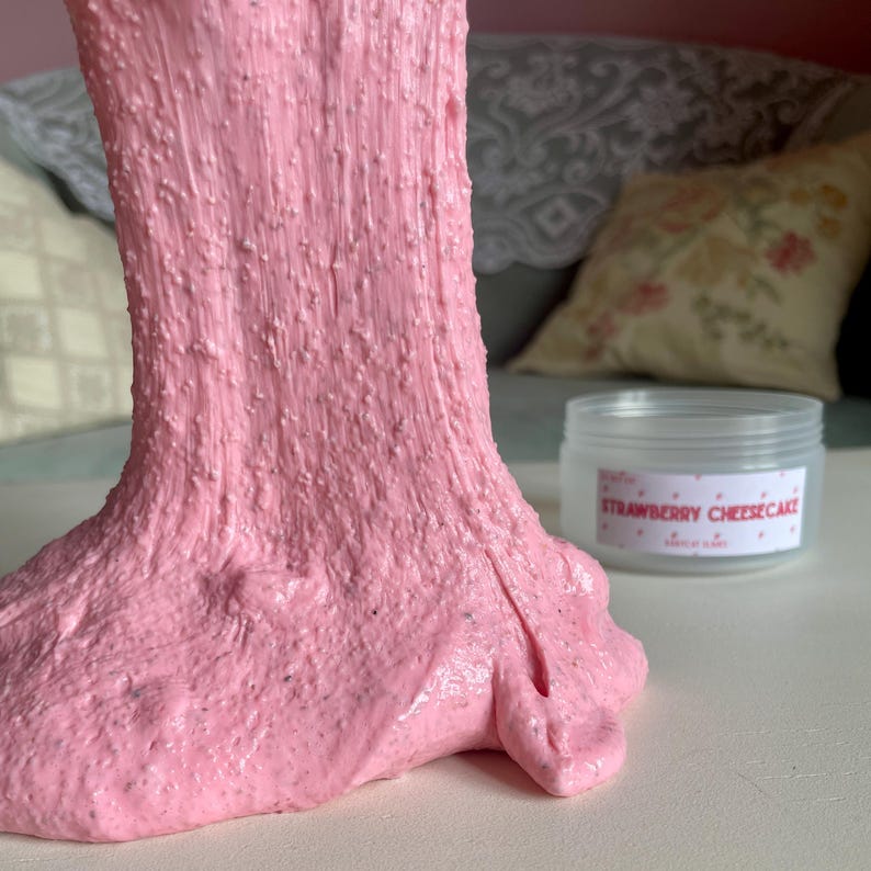 A container labeled "strawberry cheesecake by babycat slimes" is filled with a creme coloured slime with a sand crust, topped with red slime with jelly cubes and strawberry clay pieces.