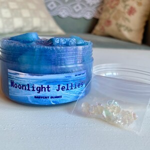 A clear, round plastic container is overflowing with blue sparkly jelly cube slime. The slime is a clear coated slime colorshifting from blue to purple, and has big squishable jelly cubes. It also has tiny opalescent jellyfish charms.