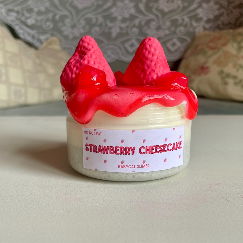 A container labeled "strawberry cheesecake by babycat slimes" is filled with a creme coloured slime with a sand crust, topped with red slime with jelly cubes and strawberry clay pieces.