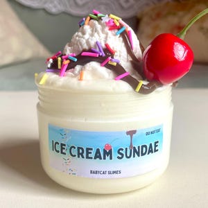 Peut inclure: Un pot de slime "Ice Cream Sundae", garni de slime blanc, de sauce au chocolat, de vermicelles colorés et d'une cerise rouge. Le pot porte une étiquette avec le nom du produit et "Babycat Slimes". Les mots "Do Not Eat" sont également visibles.