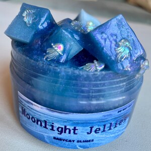 A clear, round plastic container is overflowing with blue sparkly jelly cube slime. The slime is a clear coated slime colorshifting from blue to purple, and has big squishable jelly cubes. It also has tiny opalescent jellyfish charms.