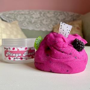 A round transparent container is full of magenta slime with tiny black seeds, resembling a pitaya dragonfruit bowl. on top are beeswax "coconut flakes" and fruit slice charms.