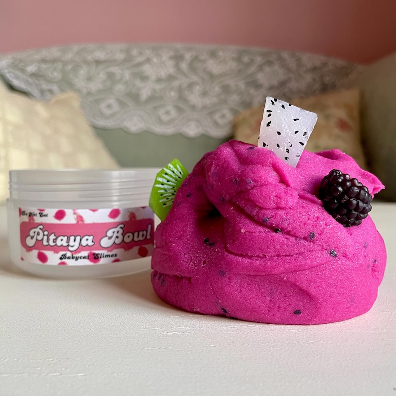A round transparent container is full of magenta slime with tiny black seeds, resembling a pitaya dragonfruit bowl. on top are beeswax "coconut flakes" and fruit slice charms.