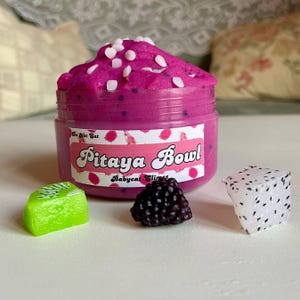 A round transparent container is full of magenta slime with tiny black seeds, resembling a pitaya dragonfruit bowl. on top are beeswax "coconut flakes" and fruit slice charms.