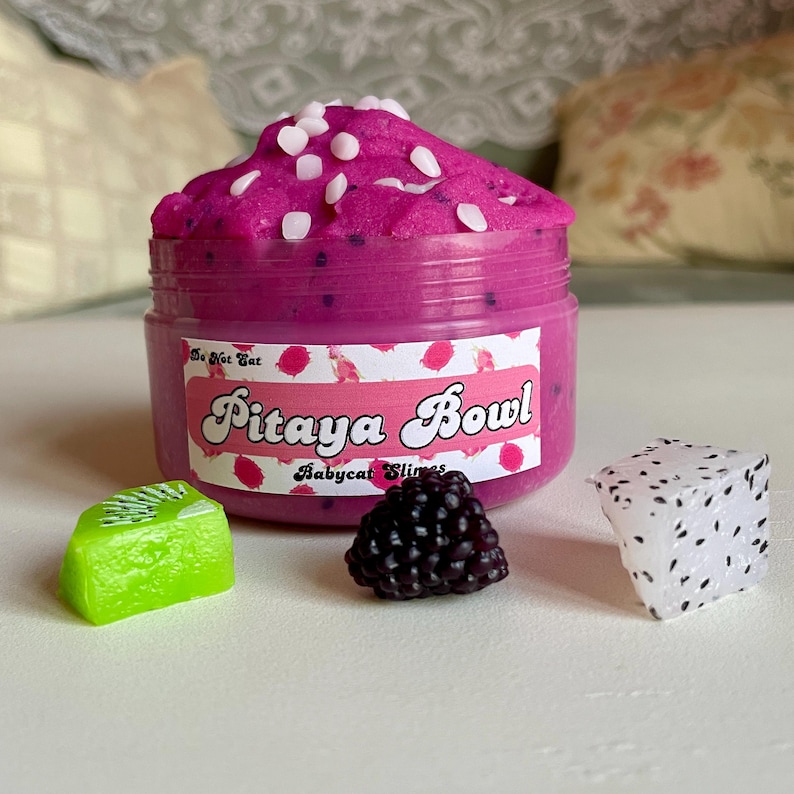A round transparent container is full of magenta slime with tiny black seeds, resembling a pitaya dragonfruit bowl. on top are beeswax "coconut flakes" and fruit slice charms.