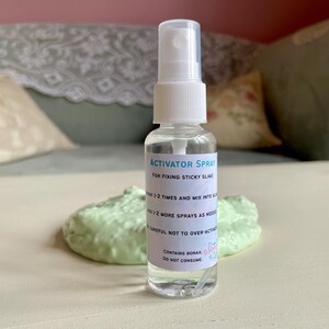 May include: A clear spray bottle labeled "ACTIVATOR SPRAY" for fixing sticky slime. The bottle has a white cap and nozzle. A blob of light green slime sits in front of the bottle. The label also says "CONTAINS BORAX. DO NOT CONSUME."