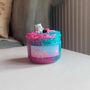 May include: A jar of colorful slime with a pink and blue glittery top, a small white bear figurine, and a label that reads "Cute but Stabby". The slime is a mix of pink and blue with glitter.