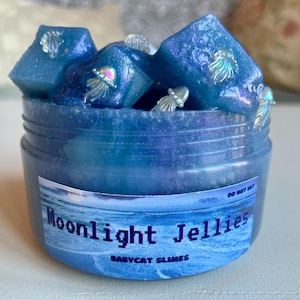 Könnte beinhalten: Ein transparentes Glas gefüllt mit blauem Schleim mit schimmerndem Glitzer und blauen, schimmernden, Plastik-Quallen-Anhängern. Das Glas hat ein Etikett mit der Aufschrift "Moonlight Jellies" und "Babycat Slimes".