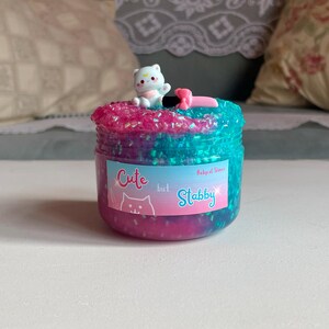 May include: A jar of colorful slime with a white cat figurine on top. The slime is a mix of pink and blue with glitter. The jar has a label that says "Cute but Stabby".