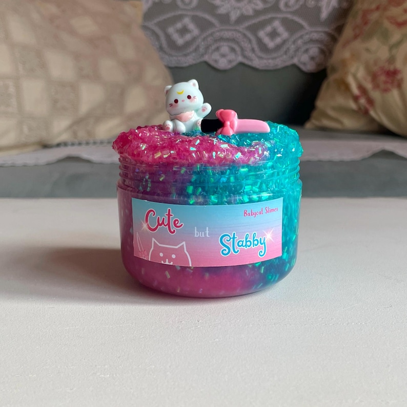 May include: A jar of colorful slime with a white cat figurine on top. The slime is a mix of pink and blue with glitter. The jar has a label that says "Cute but Stabby".