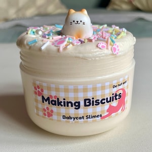 Op de afbeelding: Een potje lichtbeige slijm, bedekt met een witte, frosting-achtige substantie en kleurrijke sprinkles. Bovenop zit een klein, oranje en witte katfiguur. Het etiket leest "Making Biscuits" en "Babycat Slimes".