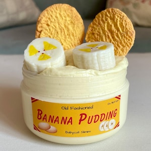 Pode incluir: Um pote de slime com tema de pudim de banana com um rótulo amarelo e vermelho que diz "Old Fashioned Banana Pudding". O slime é coberto com fatias de banana e dois biscoitos amarelos. O texto "Do Not Eat" também é visível.