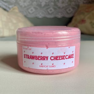 A container labeled "strawberry cheesecake by babycat slimes" is filled with a creme coloured slime with a sand crust, topped with red slime with jelly cubes and strawberry clay pieces.
