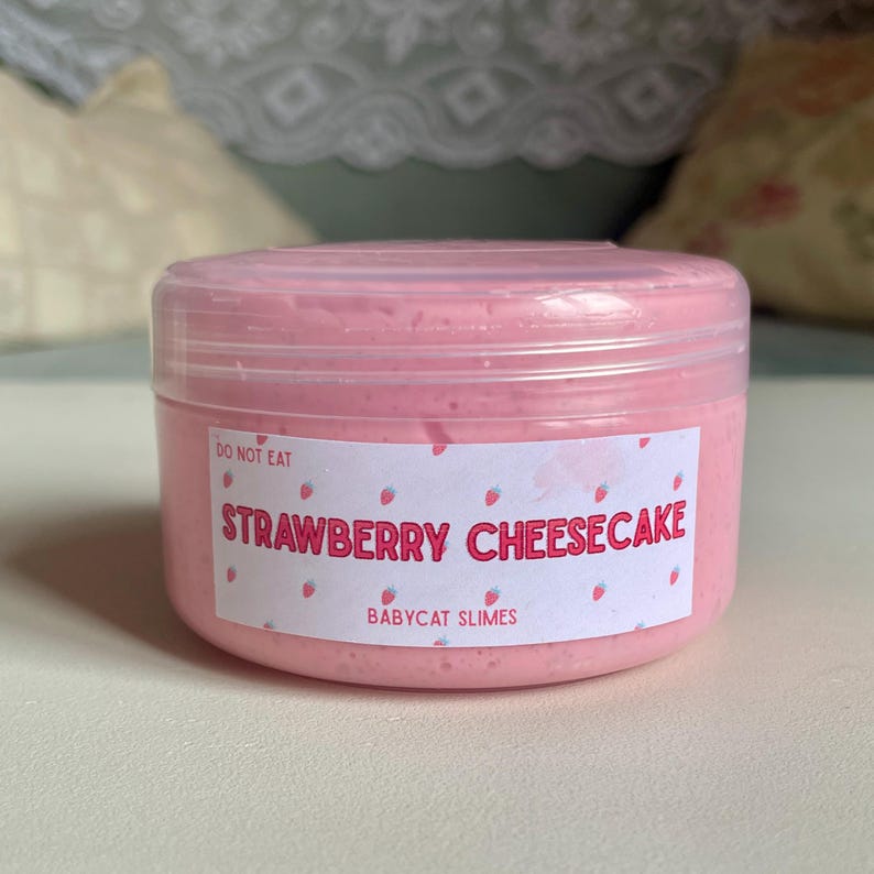 A container labeled "strawberry cheesecake by babycat slimes" is filled with a creme coloured slime with a sand crust, topped with red slime with jelly cubes and strawberry clay pieces.
