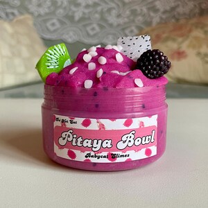 A round transparent container is full of magenta slime with tiny black seeds, resembling a pitaya dragonfruit bowl. on top are beeswax "coconut flakes" and fruit slice charms.