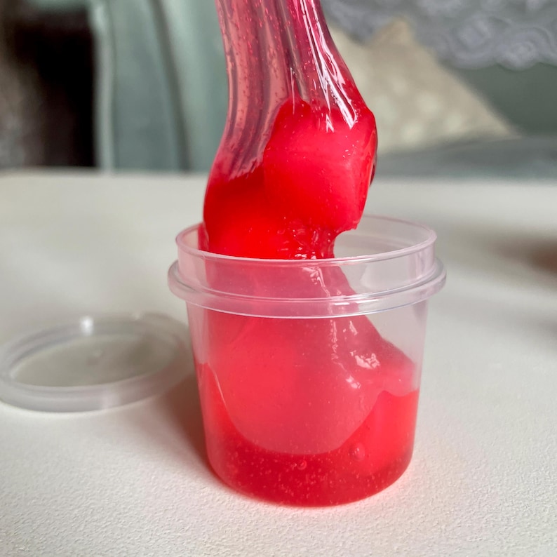 A container labeled "strawberry cheesecake by babycat slimes" is filled with a creme coloured slime with a sand crust, topped with red slime with jelly cubes and strawberry clay pieces.