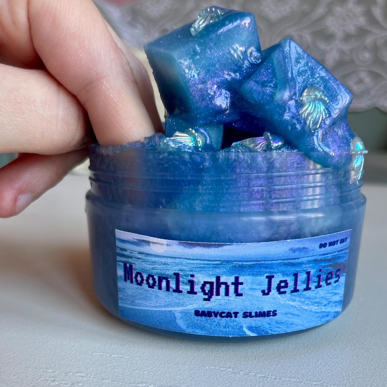 A clear, round plastic container is overflowing with blue sparkly jelly cube slime. The slime is a clear coated slime colorshifting from blue to purple, and has big squishable jelly cubes. It also has tiny opalescent jellyfish charms.