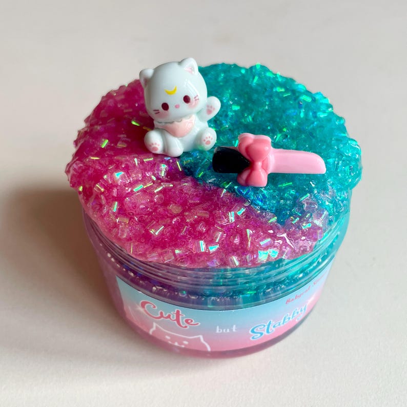 May include: A jar of slime with a split design of pink and teal glitter. A white cat figurine with a crescent moon on its forehead sits atop the pink slime. A pink and black toy knife is also on the slime. The jar has the text "Cute but Stabbin".