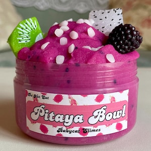 Pode incluir: Um slime rosa em forma de tigela de pitaya com granulado branco, uma fatia de kiwi, uma fatia de fruta do dragão branca e uma amora preta no topo. O slime está em um recipiente de plástico transparente com um rótulo que diz "Pitaya Bowl" e "Babycat Slimes".