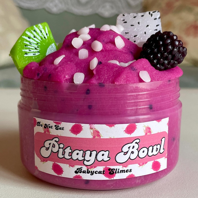 A round transparent container is full of magenta slime with tiny black seeds, resembling a pitaya dragonfruit bowl. on top are beeswax "coconut flakes" and fruit slice charms.