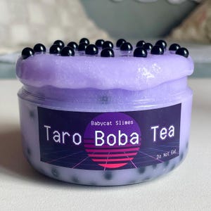 Könnte beinhalten: Ein Glas lila Schleim mit schwarzen Kugeln obenauf, beschriftet mit "Taro Boba Tea" mit dem Branding "Babycat Slimes". Das Glas hat einen transparenten Kunststoffbehälter mit einer lila, strukturierten Substanz im Inneren. Das Etikett enthält auch den Text "Do Not Eat".