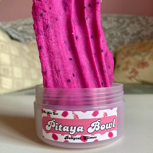 A round transparent container is full of magenta slime with tiny black seeds, resembling a pitaya dragonfruit bowl. on top are beeswax "coconut flakes" and fruit slice charms.